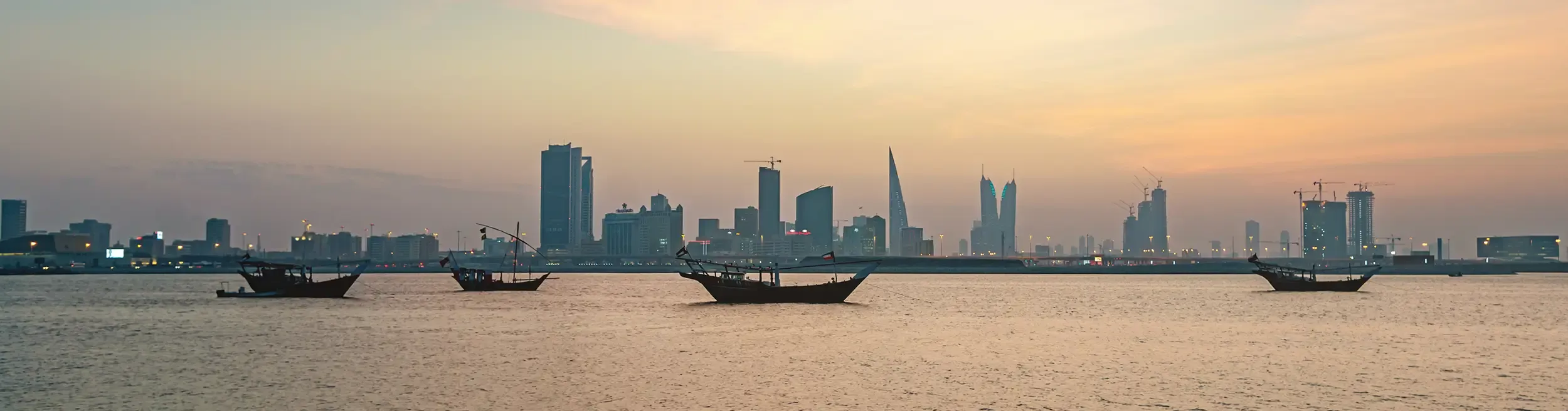 A panoramic twilight view of the Manama, Bahrain skyline, featuring traditional wooden dhow boats anchored in the foreground of the sea, contrasted against a backdrop of modern skyscrapers, luxury hotels, and the iconic Bahrain World Trade Centre.